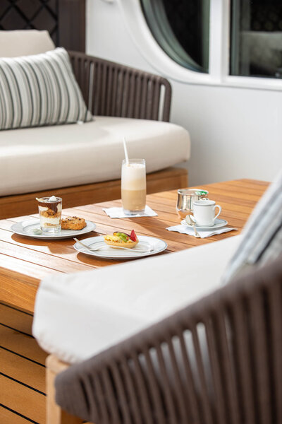 A cozy outdoor lounge area with cushioned seats and a wooden table set with coffee, desserts, and a smoothie on a yacht or cruise ship.