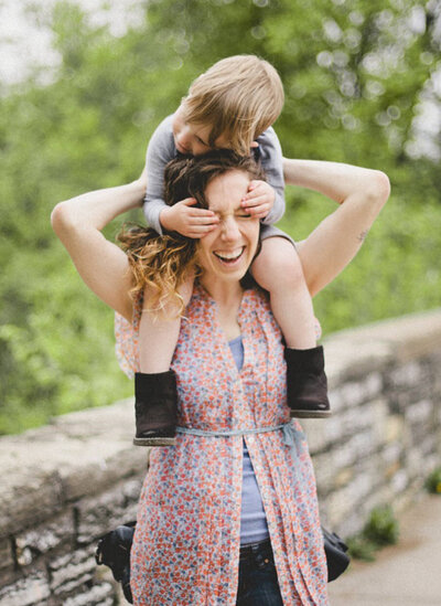 child is sitting on the shoulders of his mother and squeezing her face as she grins widely. leaves in the background