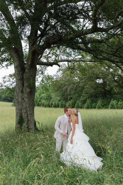 Bride and groom kiss under a tall tree in a grassy field