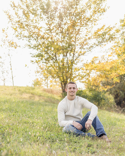 high school senior male, sitting in the grass with a tree behind him, smiling at the camera, wearing jeans and a long sleeve shirt