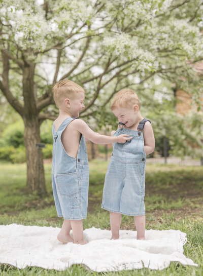 Twin boys in denim overalls playing together under spring blossoms during a lifestyle family photo session in Kansas City.