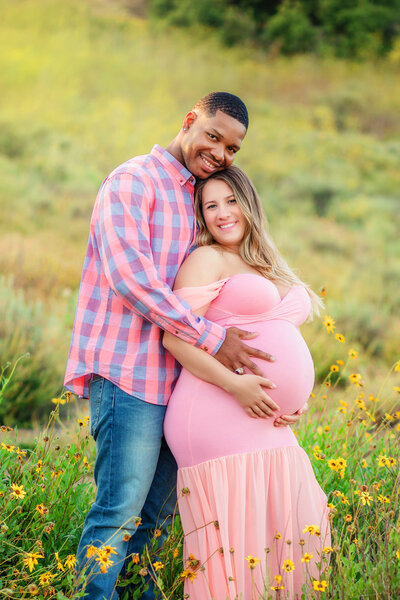 Maternity photographer, an interracial couple stand amongst flowers while a pregnant mother holds her belly
