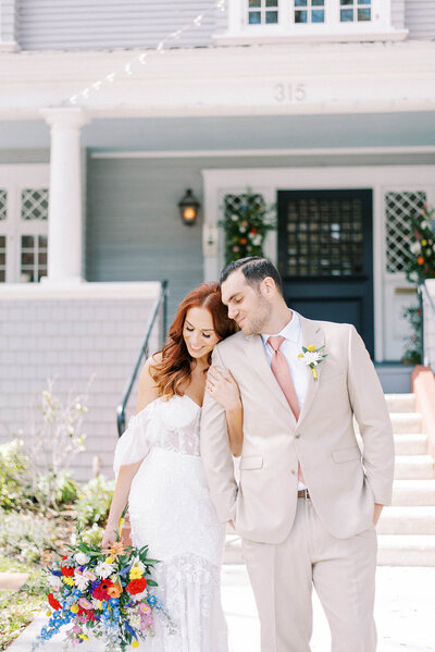 red headed bride wearing an off the shoulder lace gown while holding a brightly colored wedding bouquet and holding her groom who is wearing a tan suit in from of the Orlo