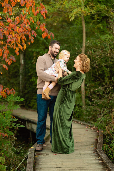 A mother and father stand in the fall foliage with their baby, laughing and tickling each other