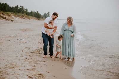 Mom, Dad, toddler and baby on Grand Bend beach for family photos at sunset. Mom is wearing a long blue dress and holding toddler's hand, Dad is holding 6 month old baby in his arms.  Mom and dad are smiling at each other and kids looking toward water for candid photo.