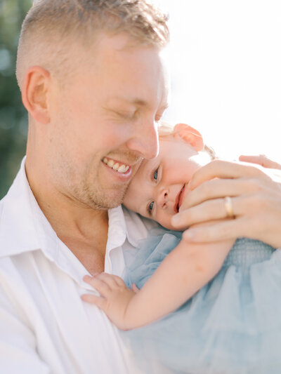 Dad in white shirt embraces one year old daughter in blue dress who is snuggling into his shoulder photographed by Little Rock photographer Bailey Feeler