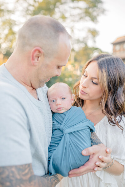 Newborn session in Westchester with mother and father lovingly holding their newborn baby. The parents gaze at their child with deep affection, surrounded by a peaceful, natural setting