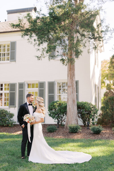 Maddie and Garrett pose for their wedding photos on the lush green lawn of Boxwood Estate. Maddie wears a stunning white wedding gown with intricate lace details, while Garrett is dressed in a sharp suit. The couple stands together, smiling and holding hands, surrounded by the estate's beautiful landscaping and tall trees. The soft, natural light adds a romantic touch to the scene, capturing a joyful and intimate moment between the newlyweds.