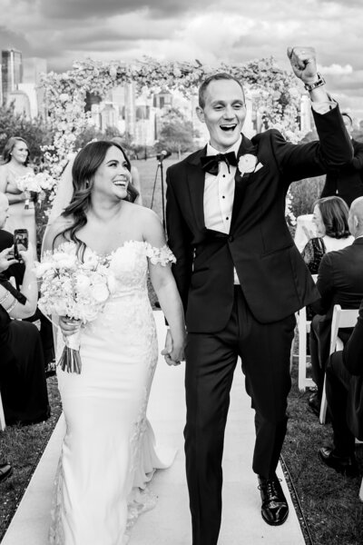 A newlywed couple walks down the aisle outdoors after their wedding ceremony at Liberty House in Jersey City.  They are holding hands and smiling. The groom raises his fist in celebration, while guests seated on either side cheer. Captured with stunning candid wedding photography, the NYC city skyline provides a breathtaking backdrop for this joyous moment.