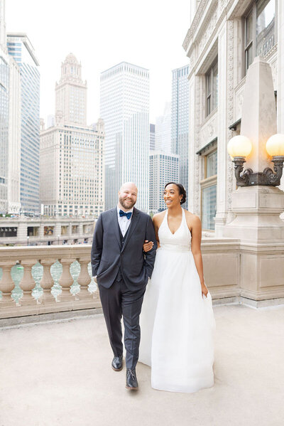 Bride and groom walk arm in arm on a city terrace, smiling and relaxed against the backdrop of Chicago’s iconic skyline. The bride wears a halter-style white gown, and the groom is in a dark suit with a blue bow tie. The scene captures a joyful moment of love and elegance amid historic architecture and riverfront views.