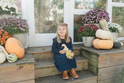 east-nashville-greenhouse-mini-sessions-by-nashville-family-photographer-dolly-delong-a-girl-holding-a-pumpkin-and-smiling-at-the-camera-for-her-fall-portraits