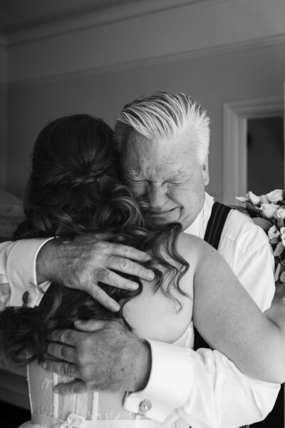 A proud father embraces his daughter holding a bouquet of flowers in a heartfelt wedding moment. Both appear emotional, standing in a softly lit room with a partially open door in the background. Black and white photo.