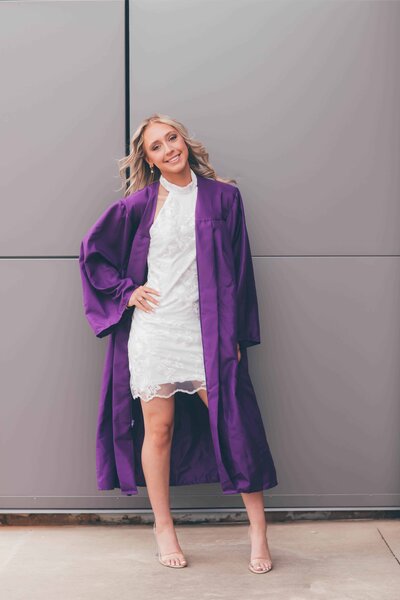A high school senior stands proudly with her hand on her hip in a white dress and purple robe celebrating graduation captured by Shreveport senior portrait photographer Britt Elizabeth Photography.