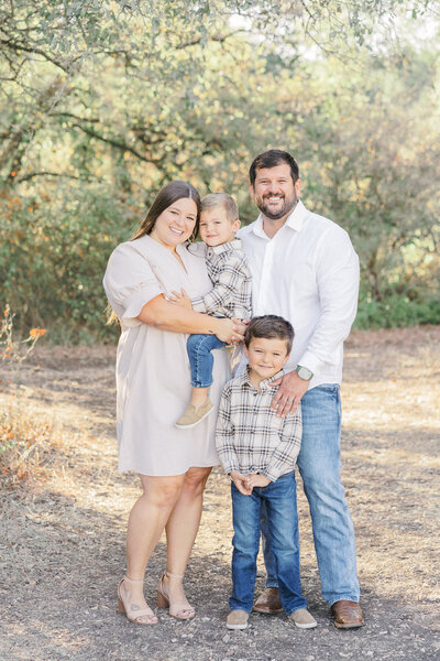 A family of four poses in a park for their family portraits in Austin, TX. They look and smile at the camera. The mom holds one of their sons in her arms while his brother stands in front of his father.