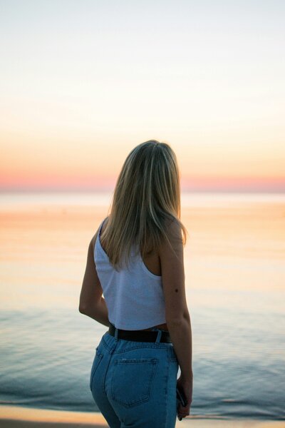 Woman standing at the shoreline looking toward the sunset, representing calm reflection and the healing process of clinical hypnosis to release subconscious patterns and restore inner peace.
