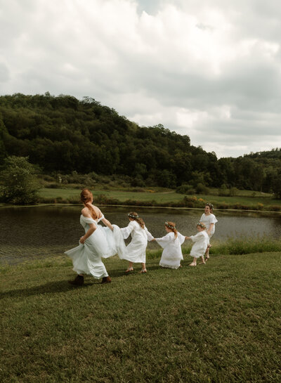Bride and bridesmaids running hand-in-hand beside a lake in the West Virginia mountains, captured by Rainstorm Photo & Video in a whimsical, Pride and Prejudice–inspired scene.