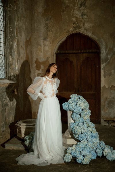 April M Payne Photography taken of a bride in a flowing white gown with a floral design, posing elegantly next to a cluster of blue hydrangeas in an atmospheric, rustic venue with vintage charm.
