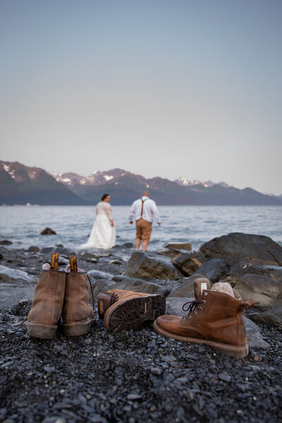 Newlyweds walk along a rocky Alaskan shoreline in wedding attire, with mountains in the background. Hiking boots rest on the beach, symbolizing adventure, wilderness, and a nature-inspired elopement.