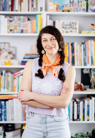 A woman with dark curly hair in pigtails with daisy earrings and a coral red bandana stands smiling with arms gently crossed. She wears a lilac colored tank top with a daisy on it and stands in front of a full wall of books in light colors.