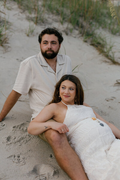 A husband and wife sit on the beach during their outer banks maternity photo session