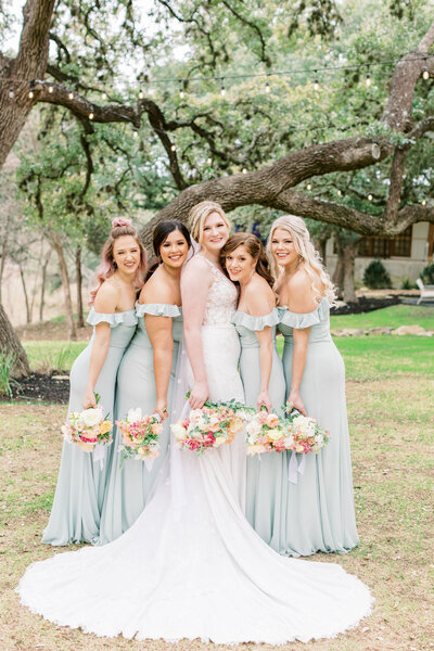 Four bridesmaids wearing the same mint green bridesmaid dresses are holding their spring colored bouquets alongside the bride in her lace wedding gown looking at the camera and smiling at the ivory oak