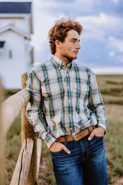 Boy in plaid shirt leans agains fence.