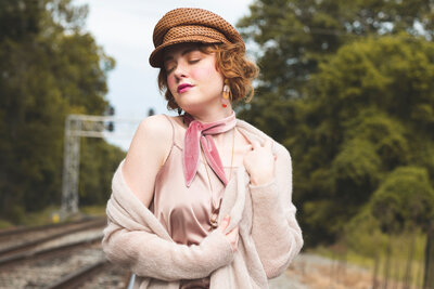 A girl sits near railroad tracks in a straw hat pink top sweather and scarf