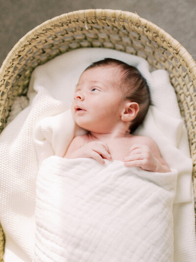 A baby wrapped in a white blanket laying on blankets in a basket and looking to the side by Katie Stansfield Photography, a Richmond newborn photographer.