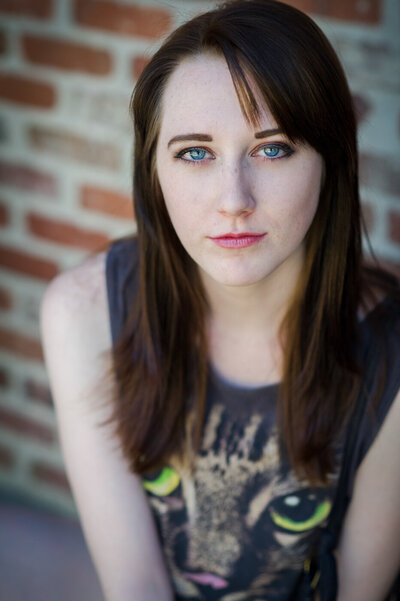Theatrical acting headshot of actress with blue eyes and brown hair wearing a cat shirt in front of brick wall.