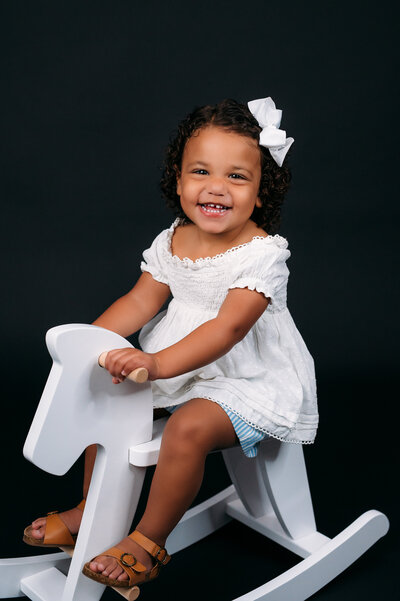 Smiling young girl sitting on a white rocking chair in a studio setting with a black backdrop