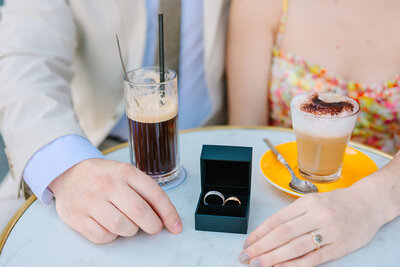 Couple at a Parisian café in the Marais near Hôtel de Sully, wearing sunglasses as the woman sips coffee during a couple’s photoshoot in Paris.