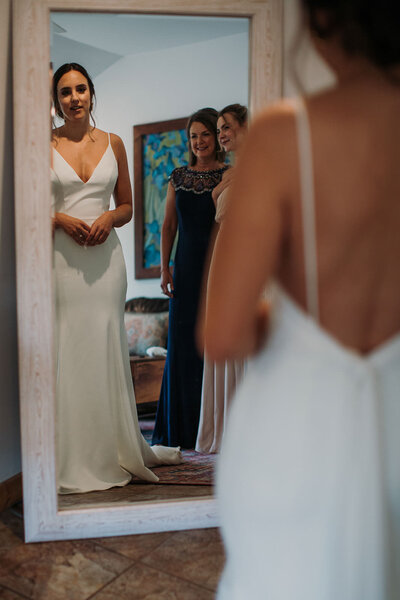A bride looking at herself in a mirror with two other people smiling behind.