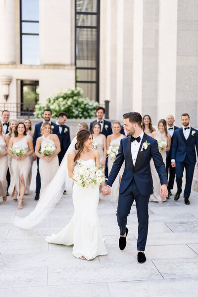 A bride and groom hold hands and smile at each other with their wedding party in the background on the landing of the Ohio Statehouse