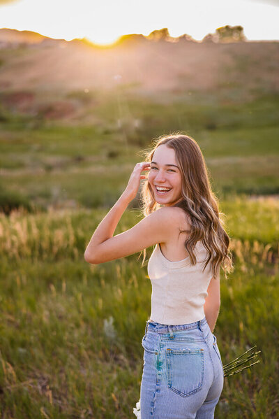 Senior girl laughs and brushes hair out of face with sun setting behind her.