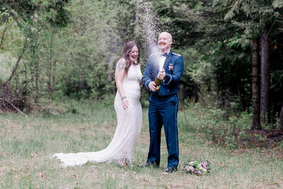 Bride in wedding dress Groom in Military Blues popping champagne in the woods.