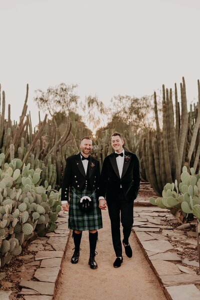 Two grooms walk hand in hand through cactus garden