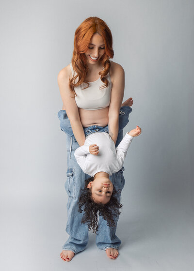 Pregnant mother in casual jeans smiling while holding toddler upside down during playful maternity photoshoot in NJ