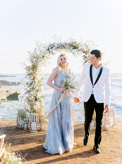 Bride and groom walking hand in hand under a floral arch at sunset, photographed by Natalie Jayne in coastal light.