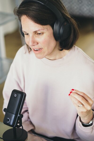Kaya wearing large headphones and a pale pink sweatshirt, speaking into a microphone at a desk.