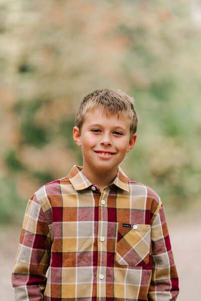 A young boy wearing a yellow and brown plaid shirt standing for his outdoor fall portrait. 