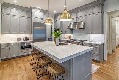 Modern kitchen interior with gray cabinets, marble island, brass pendant lights, and stainless steel appliances.