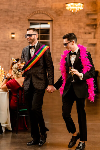a couple holding hands and smiling joyfully at a celebration, one wearing a rainbow pride sash and the other a pink feather boa, symbolizing LGBTQ+ pride and love.