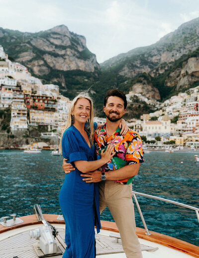 man and woman standing on a boat in the water with mountains in the background