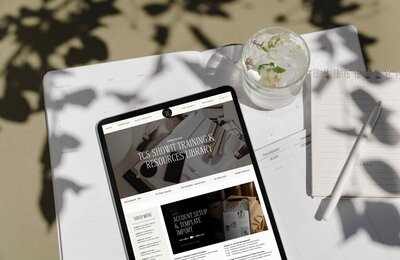 Tablet displaying The Closers Society Showit training library dashboard on a flat lay with a planner, pen, and floral water glass in natural light