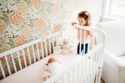 Toddler peers over the crib rails at her new baby sister during In-Home Newborn Photoshoot in Charleston, SC.