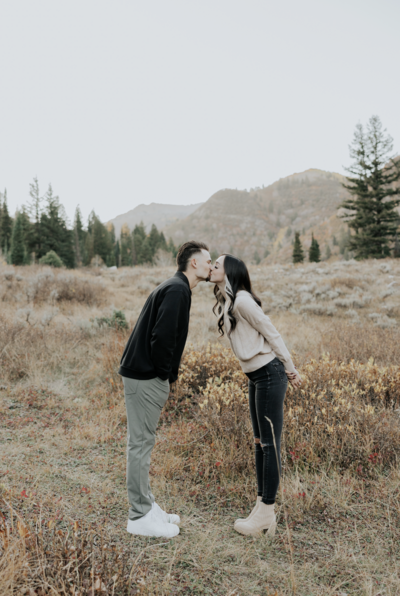 Couple lean forward and kiss while standing in mountains and field.