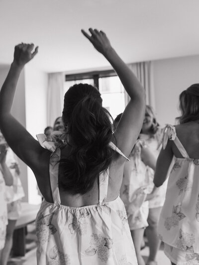 A bridal party in floral dresses joyously dance in a sunlit room. The scene conveys a sense of celebration and happiness, arms raised in excitement.