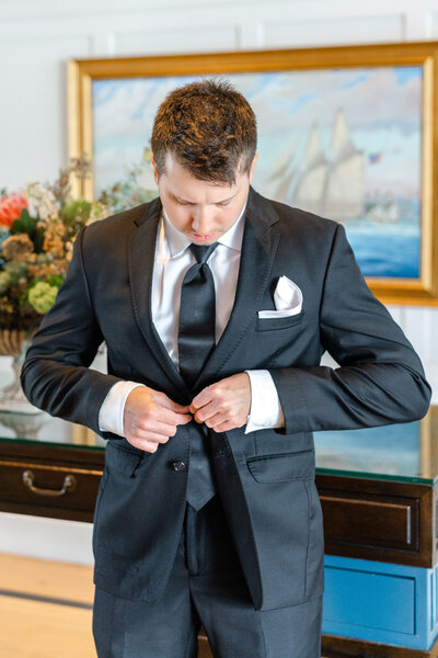 Groom buttoning black suit jacket at Lido House before wedding, with nautical artwork in the background