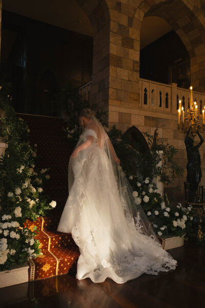 bride with long flowing lacy gown walking up a dramatic set of stairs surrounded by flowers at dover hall in richmond virginia