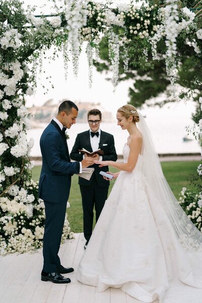 Bride and groom exchanging vows during a waterfront wedding ceremony under a stunning floral arch, with an officiant standing between them